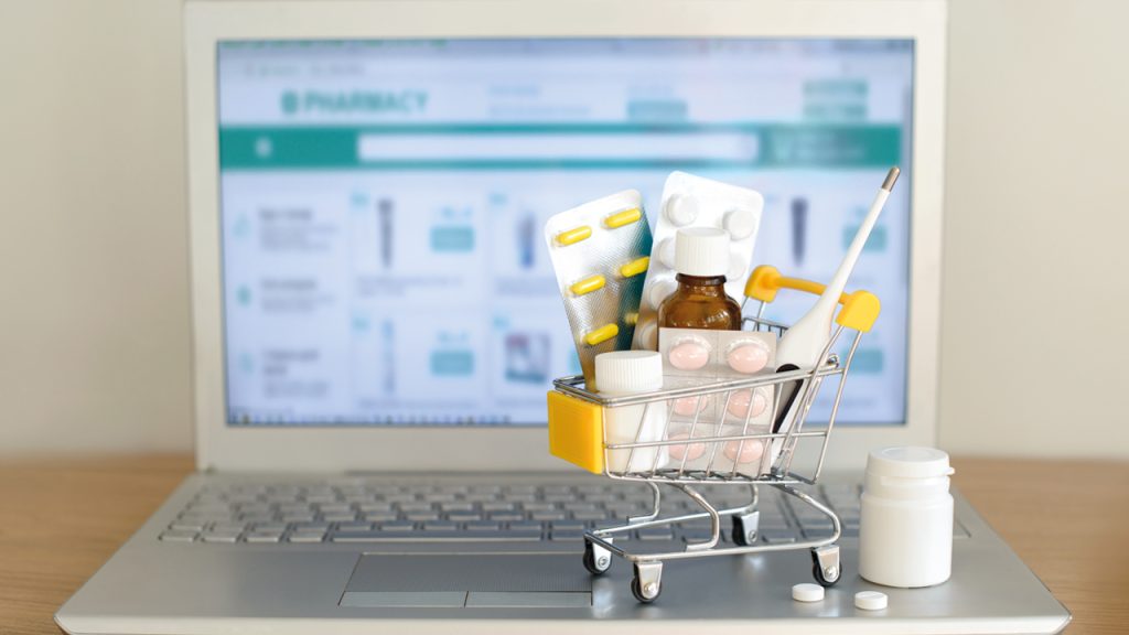 Shopping cart toy with medicaments in front of laptop screen with pharmacy web site on it. Pills, blister packs, medical bottles, thermometer set. Health care and internet shopping.